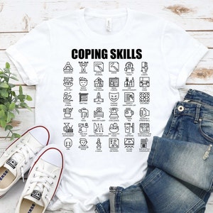 May include: White t-shirt with the text "Coping Skills" printed in black. The shirt features a grid of 36 black and white icons representing different coping skills, such as meditating, exercising, writing in a journal, drawing, listening to music, taking a bath, playing with a pet, enjoying nature, cleaning the house, reading a book, using aromatherapy, playing a game, cooking a meal, knitting, praying, finding humor, making a gratitude list, coloring, doing yoga, getting enough sleep, accepting a challenge, practicing deep breathing, drinking tea, squeezing a stress ball, playing a musical instrument, asking for help, eating healthy food, limiting caffeine, scheduling time for yourself, crying, smiling, taking a walk, doodling on paper, going to see a movie, and eating a little chocolate.