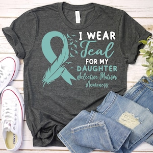 May include: Teal ribbon with a feather design on a gray t-shirt. The shirt reads "I wear teal for my daughter Selective Mutism Awareness".