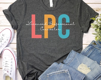 以下が含まれることがあります： 濃いグレーのTシャツに「LPC Licensed Professional Counselor」という文字がカラフルな文字で書かれています。文字はピンク、オレンジ、ブルーです。