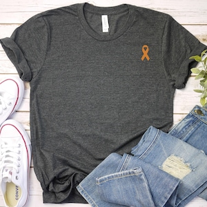 May include: A dark gray t-shirt with a rolled up sleeve and an orange ribbon on the left chest. The t-shirt is laying on a white wooden surface with a pair of blue jeans and white sneakers.