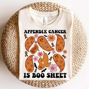 May include: A white t-shirt with a graphic design of ghosts, bats, and flowers. The text "Appendix Cancer is Boo Sheet" is printed in black letters.