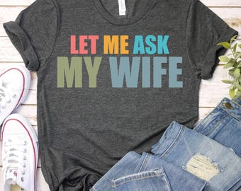 May include: Dark gray t-shirt with the text "LET ME ASK MY WIFE" in colorful block letters. The shirt is paired with blue jeans and white canvas sneakers. A casual, everyday wear tee.
