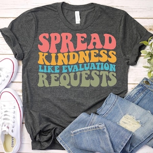 May include: A dark grey t-shirt featuring the phrase "SPREAD KINDNESS LIKE EVALUATION REQUESTS" in retro colours: coral, orange, teal, and olive green. White canvas trainers and blue jeans are also visible in the image.