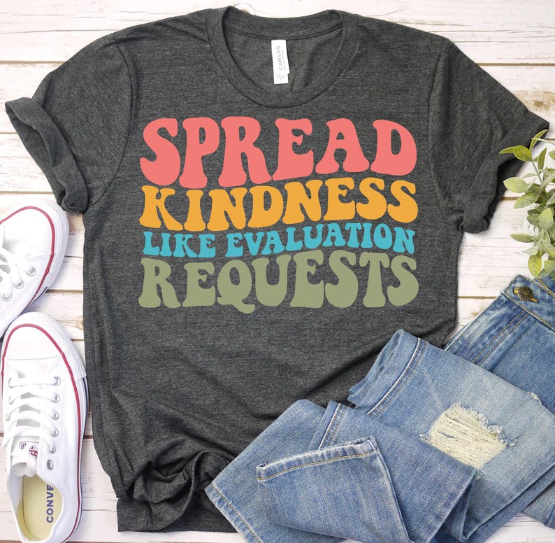 May include: A dark grey t-shirt featuring the phrase "SPREAD KINDNESS LIKE EVALUATION REQUESTS" in retro colours: coral, orange, teal, and olive green. White canvas trainers and blue jeans are also visible in the image.