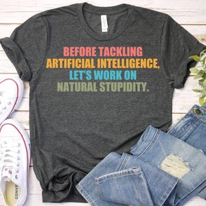 May include: Dark gray t-shirt with the text "BEFORE TACKLING ARTIFICIAL INTELLIGENCE, LET'S WORK ON NATURAL STUPIDITY." The text is in various colors. White canvas sneakers and blue jeans are also in the image.
