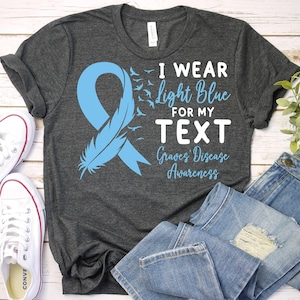 May include: A dark grey t-shirt with a light blue ribbon and feather design. The text on the shirt reads "I wear light blue for my TEXT Graves' Disease Awareness".