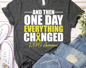 Puede incluir: Camiseta gris con texto blanco que dice "And then one day everything changed DIPG Awareness". Una cinta amarilla está incorporada en la palabra "changed".