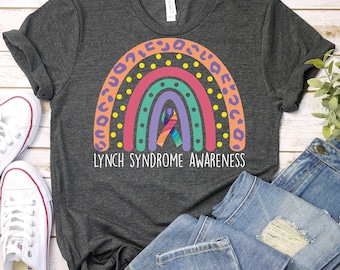 Puede incluir: Camiseta gris oscuro con un diseño de arcoíris colorido y el texto "LYNCH SYNDROME AWARENESS". El arcoíris presenta estampado de leopardo naranja, puntos amarillos y una cinta morada. Zapatillas blancas y vaqueros azules también son visibles.