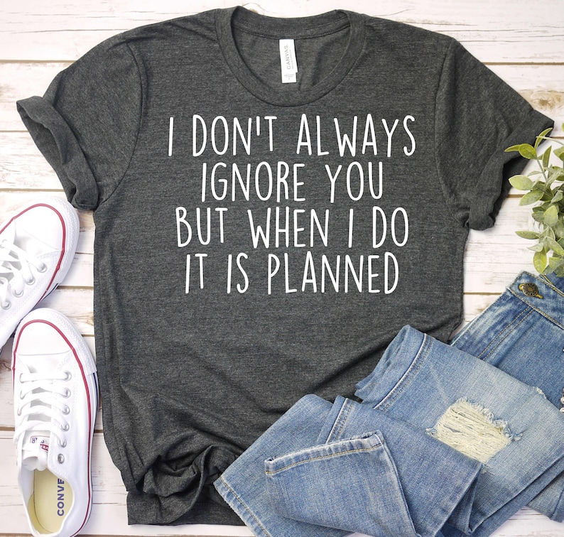 May include: Dark gray t-shirt with the text "I DON'T ALWAYS IGNORE YOU BUT WHEN I DO IT IS PLANNED" in white. The shirt is paired with blue jeans and white canvas sneakers.