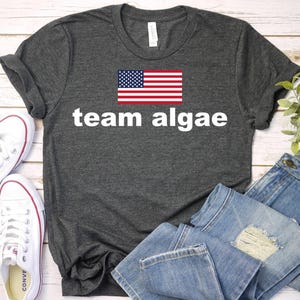 May include: A dark heather gray short-sleeve t-shirt featuring a graphic of the United States flag centered above the white text "team algae" in a bold, sans-serif font. The shirt has a crew neckline and a standard fit.