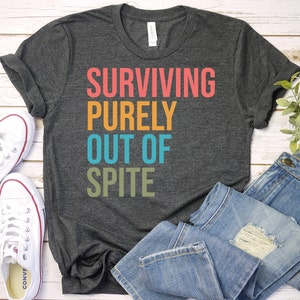 Pu&ograve; includere: T-shirt grigio scuro con la scritta "SURVIVING PURELY OUT OF SPITE" in corallo, arancione, turchese e verde oliva. La maglietta &egrave; appoggiata con scarpe da ginnastica bianche e jeans blu.