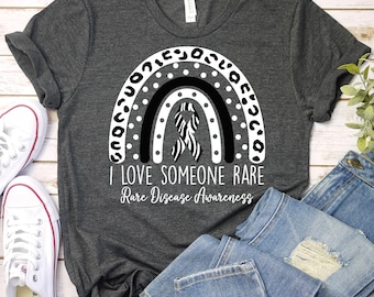 May include: A gray t-shirt with a black and white rainbow design and a zebra print ribbon. The text "I love someone rare" and "Rare Disease Awareness" is printed below the rainbow.