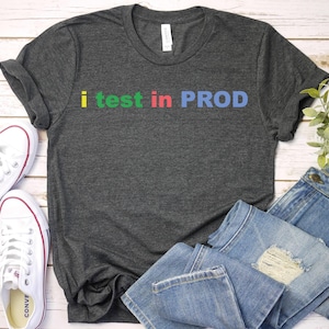May include: Dark gray t-shirt with the text "i test in PROD" in colorful letters. The shirt is paired with white sneakers and blue jeans, creating a casual and comfortable look. The shirt is made of a soft material.