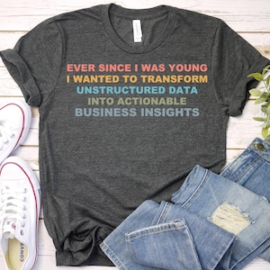 May include: Dark gray t-shirt with the text "EVER SINCE I WAS YOUNG I WANTED TO TRANSFORM UNSTRUCTURED DATA INTO ACTIONABLE BUSINESS INSIGHTS" in colorful lettering. White sneakers and blue jeans are also in the image.