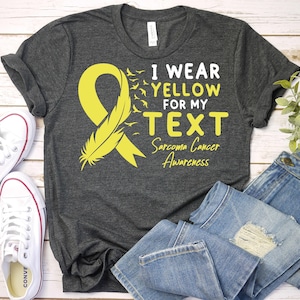 May include: A gray t-shirt with a yellow ribbon and feather design. The text on the shirt reads "I wear yellow for my TEXT Sarcoma Cancer Awareness".