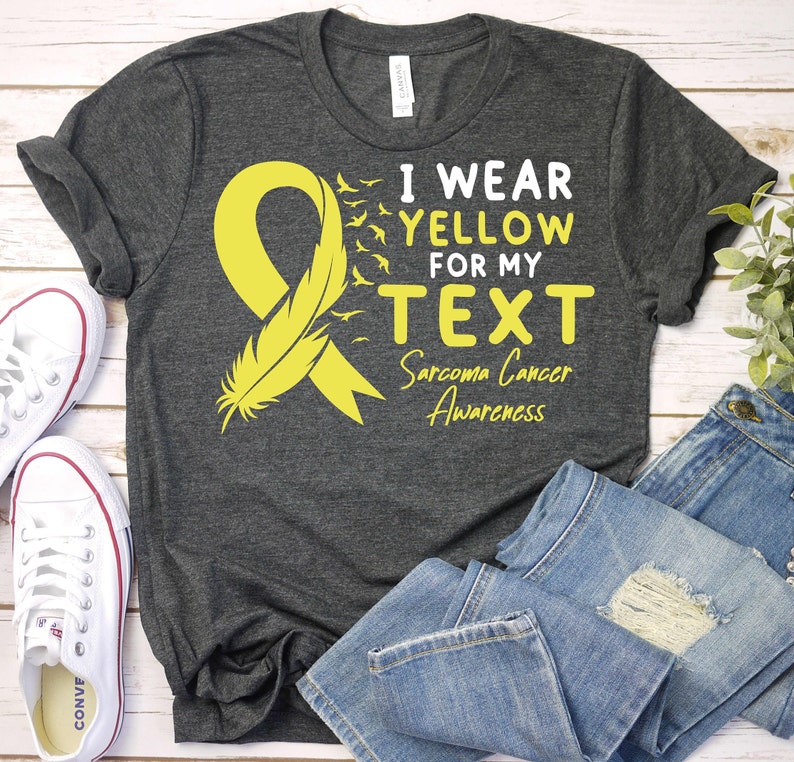 May include: A gray t-shirt with a yellow ribbon and feather design. The text on the shirt reads "I wear yellow for my TEXT Sarcoma Cancer Awareness".