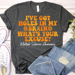 May include: Dark gray t-shirt with the text "I'VE GOT HOLES IN MY BRAIN WHAT'S YOUR EXCUSE? Multiple Sclerosis Awareness" in orange. The shirt is paired with blue jeans and white sneakers.
