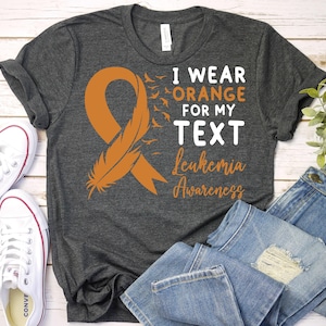 May include: A dark grey t-shirt with an orange ribbon and a feather design. The text on the shirt reads "I wear orange for my text leukemia awareness".