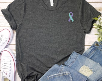 May include: A dark heather grey t-shirt with a teal and purple ribbon on the left chest. The shirt is laying flat on a white wooden surface with a pair of white sneakers and a pair of blue jeans.