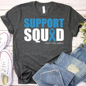 May include: A dark gray t-shirt with the text "SUPPORT SQUAD" in white letters with a blue ribbon. The text "#BEHCET'S DISEASE AWARENESS" is printed in smaller letters below the ribbon.