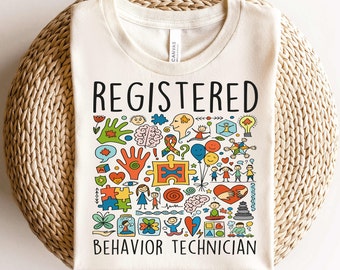 May include: A beige t-shirt with the text "REGISTERED BEHAVIOR TECHNICIAN" printed on the front. The text is surrounded by colorful illustrations of children, hearts, puzzle pieces, and other symbols.