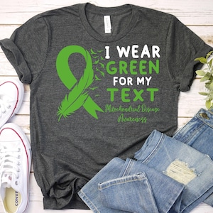 May include: A dark gray t-shirt with a green ribbon and a feather design. The text on the shirt reads "I wear green for my TEXT Mitochondrial Disease Awareness".