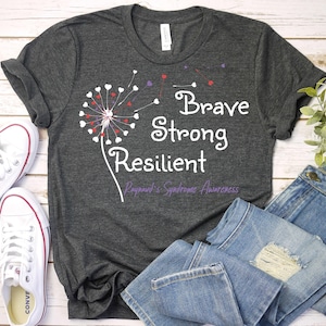 May include: A dark gray t-shirt with a white dandelion design and the words "Brave Strong Resilient" in white. The text "Raynaud's Syndrome Awareness" is in purple below the dandelion.