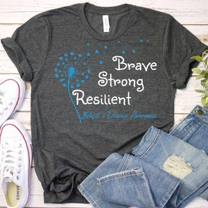 May include: A dark gray t-shirt with a blue dandelion design and the text "Brave Strong Resilient Behcet's Disease Awareness".