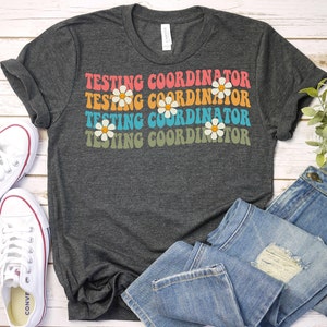 May include: Dark gray t-shirt with the words "TESTING COORDINATOR" in a retro font, with daisy graphics. The shirt is paired with white sneakers and blue jeans, creating a casual, everyday look.