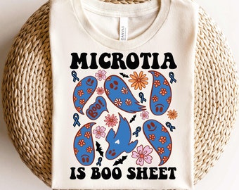 May include: Cream-colored t-shirt with the words "MICROTIAS IS BOO SHEET" in black. The shirt features blue paisley-shaped ghosts with orange and pink flowers, blue ribbons, and black bats. The shirt is folded on a woven surface.