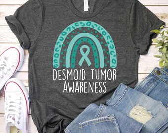 Puede incluir: Una camiseta gris oscuro con un arcoíris azul turquesa y un lazo. El texto "Desmoid Tumor Awareness" está impreso debajo del arcoíris.