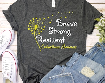 May include: Dark gray t-shirt with the words "Brave, Strong, Resilient, Endometriosis Awareness" in white and yellow. A yellow dandelion design is on the left side of the shirt. White sneakers and blue jeans are in the background.