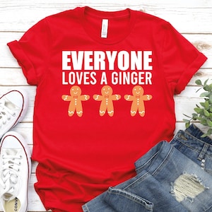 May include: A red t-shirt with the text "EVERYONE LOVES A GINGER" in white block letters. Below the text are three gingerbread men with orange bodies, white accents, and bow ties. The shirt is folded with white sneakers and blue jeans.