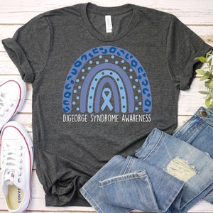 May include: A dark gray t-shirt with a blue rainbow design and a blue ribbon. The text "DiGeorge Syndrome Awareness" is printed below the rainbow.