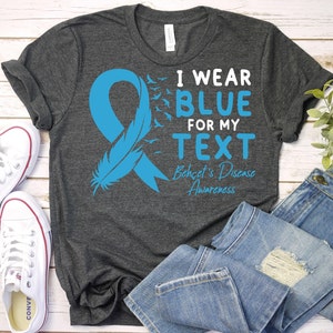 May include: A dark gray t-shirt with a blue ribbon, a feather, and flying birds. The text on the shirt reads "I wear blue for my text Behcet's Disease Awareness".