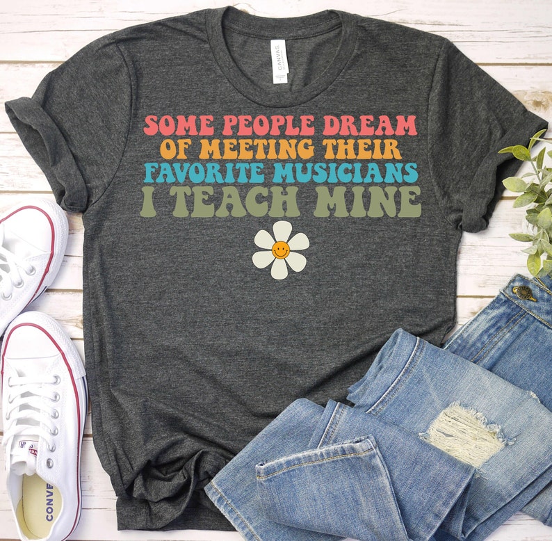 May include: A dark grey t-shirt with the text "Some people dream of meeting their favorite musicians I teach mine" in rainbow colors. A yellow daisy with a smiley face is below the text.