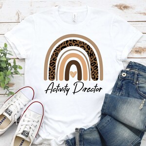 May include: White t-shirt with a brown, orange, and black leopard print rainbow design and the text "Activity Director".