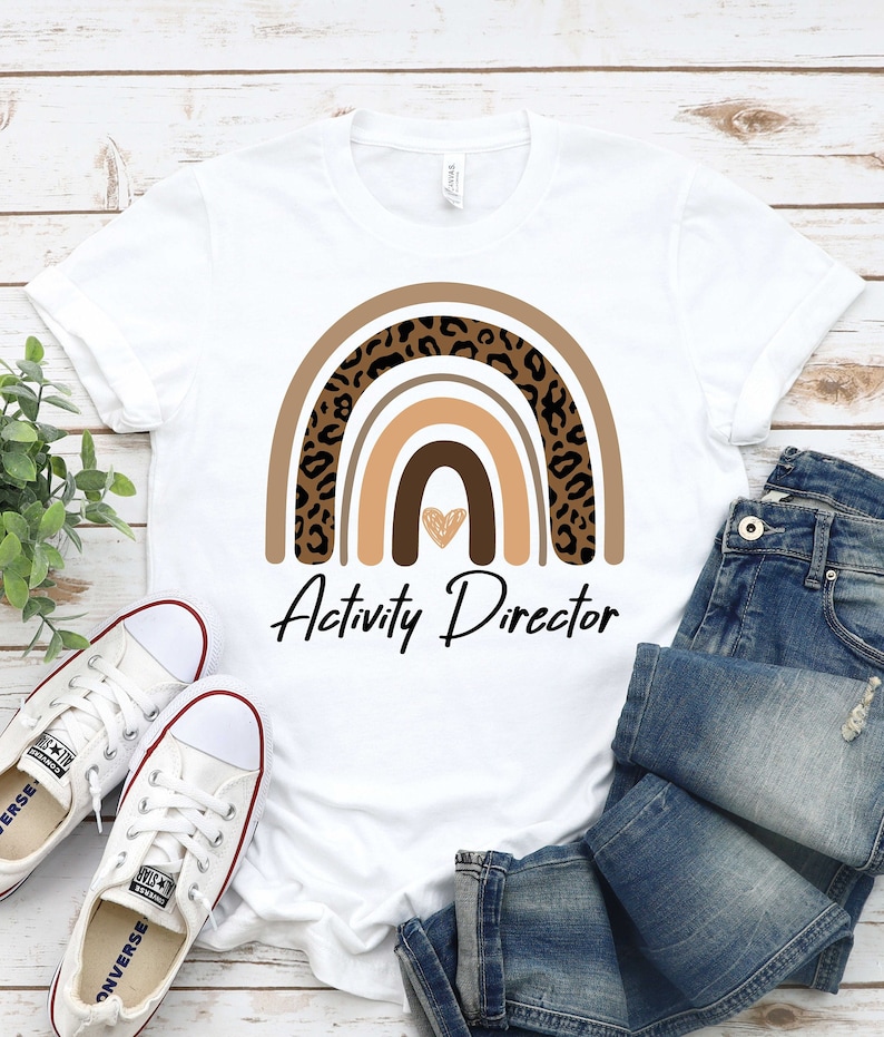 May include: White t-shirt with a brown, orange, and black leopard print rainbow design and the text "Activity Director".
