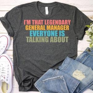 May include: A dark gray t-shirt with the text "I'm that legendary general manager everyone is talking about" in colorful letters.