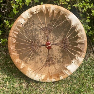 May include: A round, handcrafted drum with a natural hide surface. The drumhead is stretched over a wooden frame and secured with a woven pattern. A red stone is at the center of the drum.