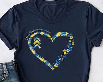 Può includere: T-shirt blu navy con un design a forma di cuore nei colori blu e giallo, con un motivo marmorizzato e impronte di mani. Il testo "Down Syndrome Awareness" è sopra il cuore. La maglietta è mostrata con pantaloncini di jeans.
