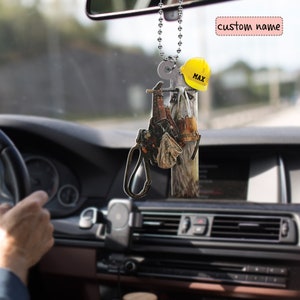 Könnte beinhalten: Ein Rückspiegel-Anhänger mit einem Holz-Strommast mit einem gelben Helm, einem Werkzeuggürtel und einem Paar Handschuhen. Der Helm trägt den Namen "MAX".