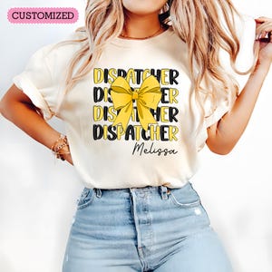 Puede incluir: Una camiseta blanca con un lazo amarillo y el texto "DISPATCHER" repetido cuatro veces. El nombre "Melissa" está impreso debajo del texto.