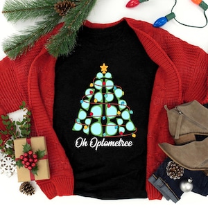 May include: A black t-shirt with a green Christmas tree design and the text "Oh Optometree". The tree is decorated with colorful lights and has a star on top. The shirt is laid out on a red knitted cardigan and blue jeans.