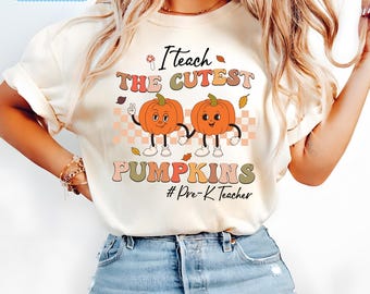 Op de afbeelding: Een wit T-shirt met de tekst "Ik leer de schattigste pompoenen #Pre-K Teacher" in kleurrijke letters. Twee cartoon-pompoenen met gezichten houden elkaars hand vast.