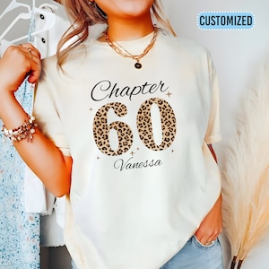 May include: A white t-shirt with the text "Chapter 60" in black with a leopard print design. The name "Vanessa" is printed below the number.