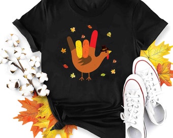 Może przedstawiać: Czarny T-shirt z grafiką brązowego indyka. Indyk wykonuje znak "Kocham cię" w języku migowym. Indyk nosi kapelusz pielgrzyma, a wokół niego znajdują się jesienne liście.