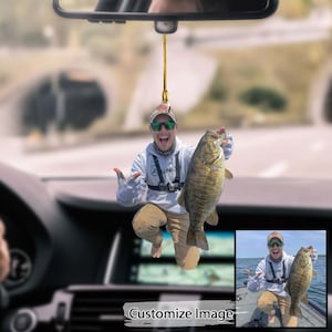 Könnte beinhalten: Ein Auto-Lufterfrischer mit einem Mann, der einen großen Barsch hält. Der Mann trägt ein weißes Hemd, eine Khakihose und eine Sonnenbrille. Der Fisch ist braun und grün mit schwarzen Flecken. Der Lufterfrischer hängt am Rückspiegel eines Autos.