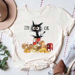 May include: A white t-shirt with a black cat illustration and the text "I'M OK". The cat is surrounded by boxes and envelopes, including a red mailbox.