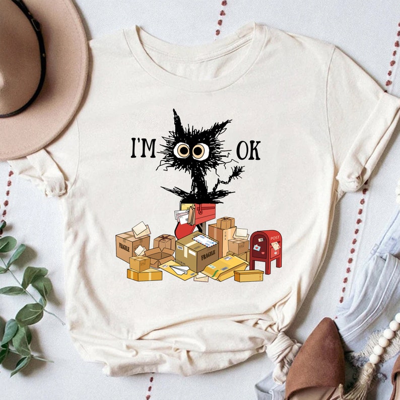 May include: A white t-shirt with a black cat illustration and the text "I'M OK". The cat is surrounded by boxes and envelopes, including a red mailbox.